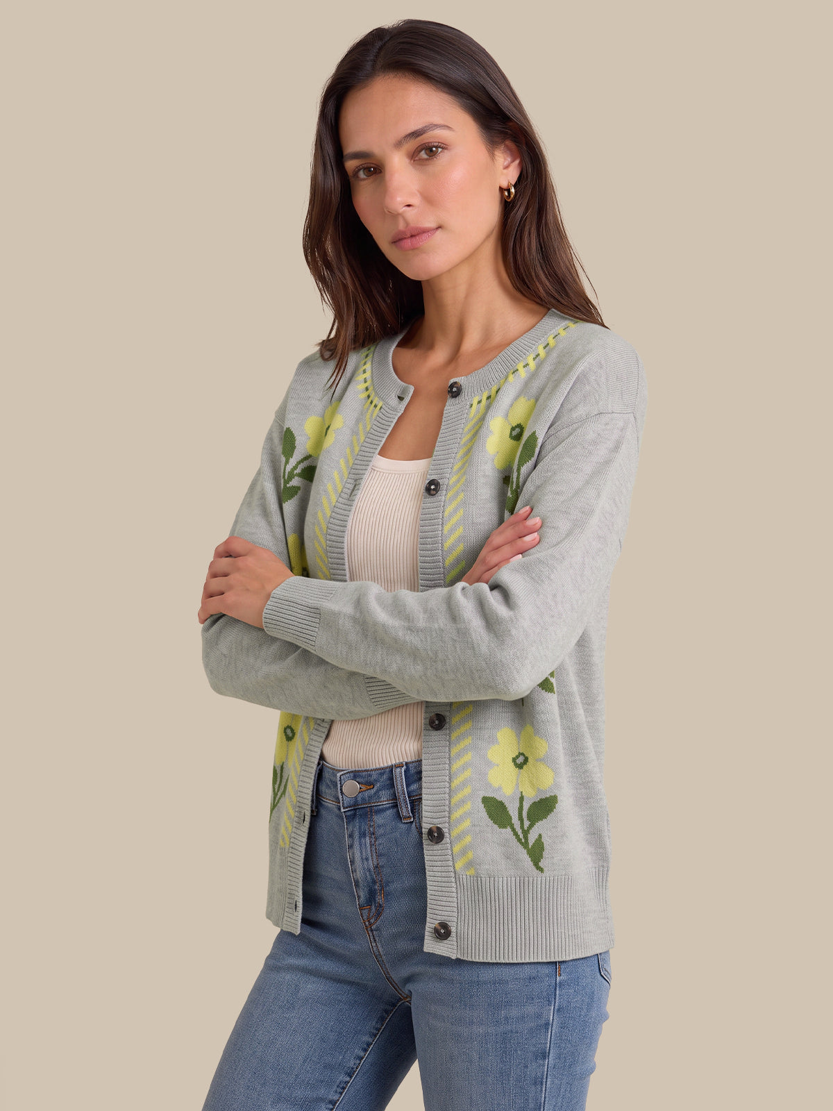 A woman modeling a light gray knit cardigan with yellow and green floral embroidery, worn over a cream tank top and blue jeans against a solid beige background.