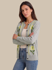 A woman modeling a light gray knit cardigan with yellow and green floral embroidery, worn over a cream tank top and blue jeans against a solid beige background.