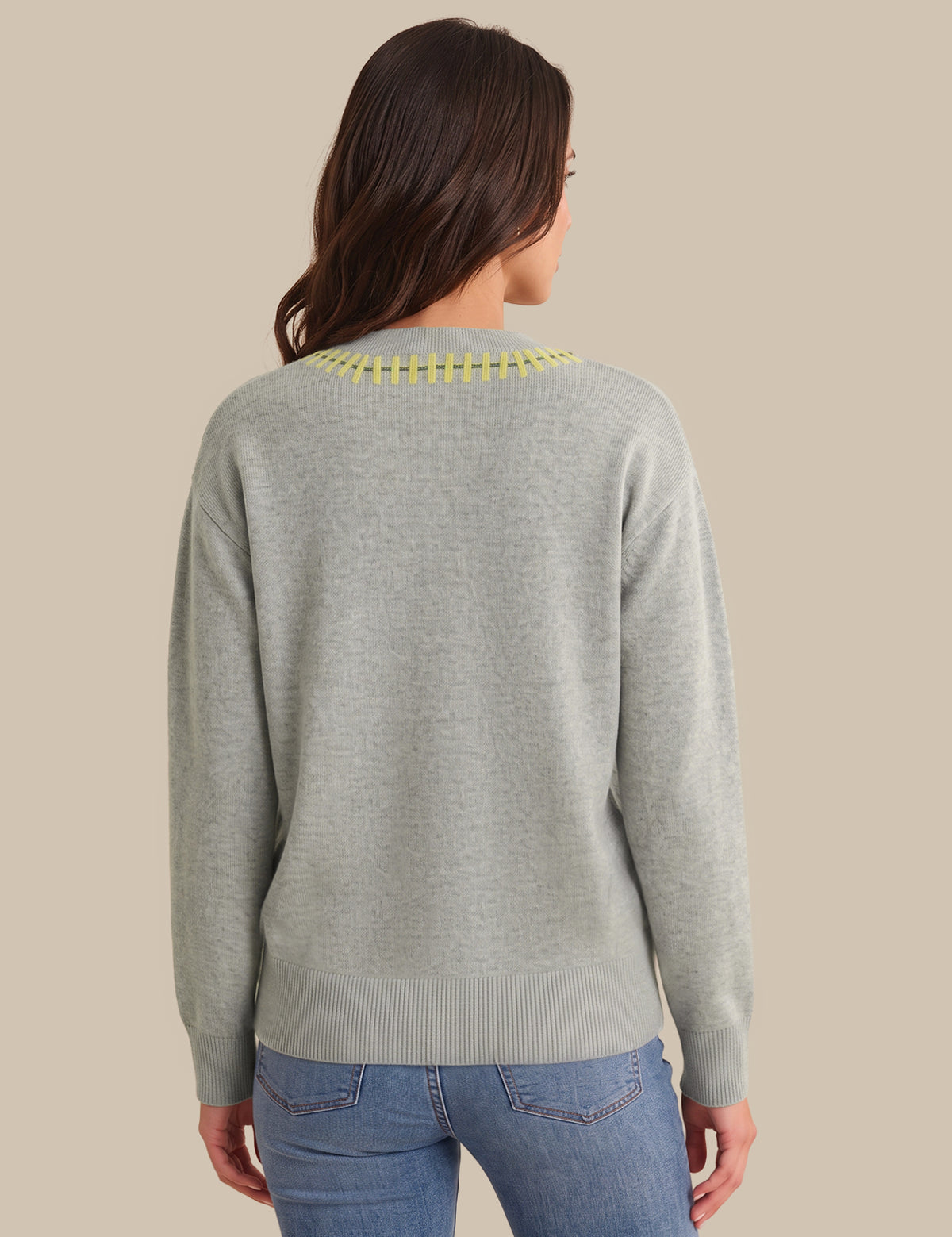 A woman viewed from the back wearing a light gray knit sweater with a yellow embroidered collar and blue jeans, set against a solid beige background.