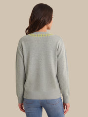 A woman viewed from the back wearing a light gray knit sweater with a yellow embroidered collar and blue jeans, set against a solid beige background.