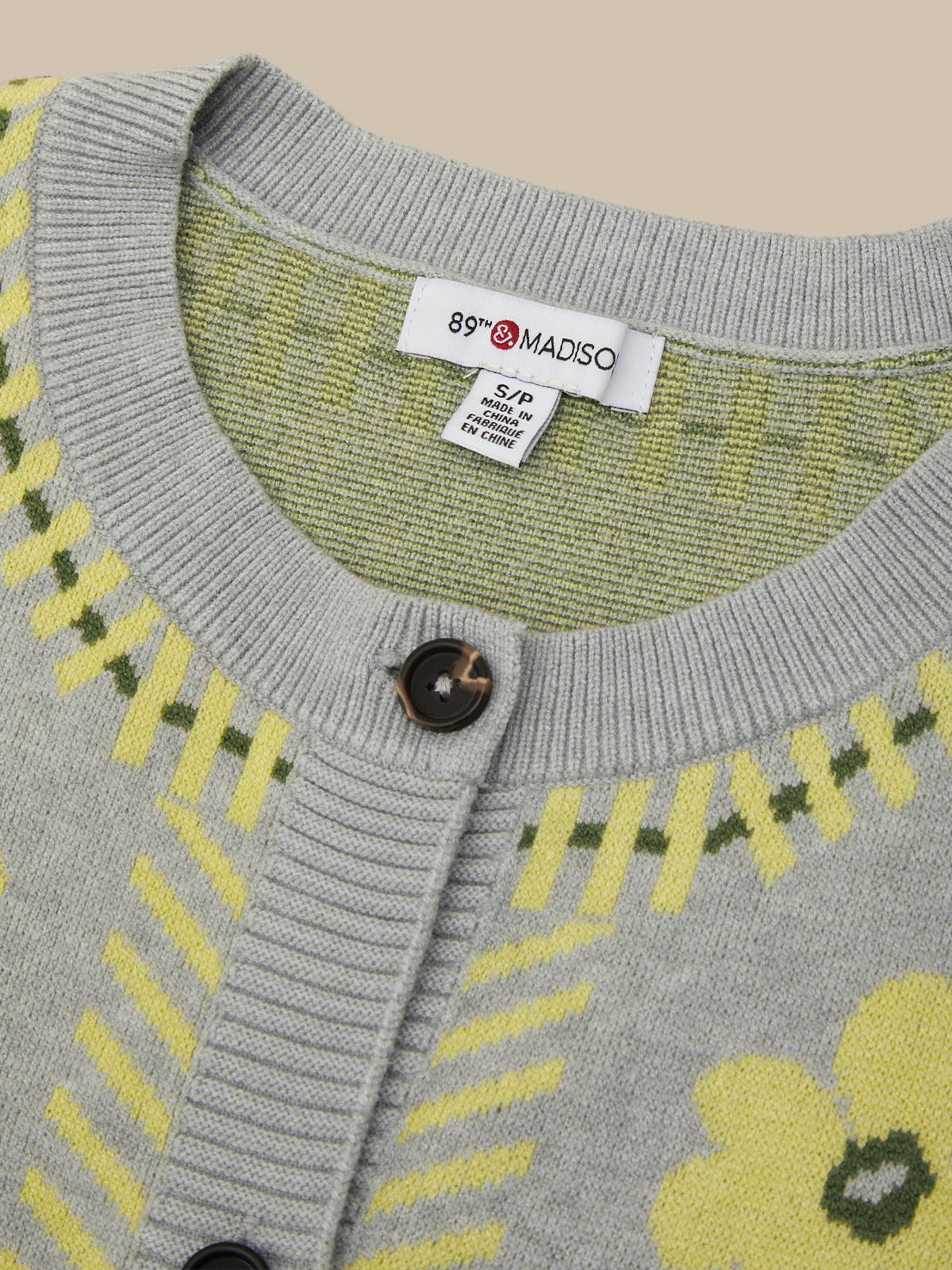 Close-up of a gray knit cardigan sweater with yellow and green geometric and floral patterns, featuring a white tag with the brand name '89 MADISO' inside the collar.