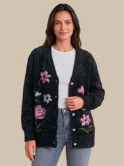 A woman modeling a black fuzzy open-front cardigan with pink, blue, and green floral embroidery and gold-tone buttons, worn over a white top and blue jeans against a solid beige background.