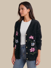 A woman wearing a black fuzzy open-front cardigan with pink, blue, and green floral embroidery and gold-tone buttons, styled over a white top and blue jeans against a solid beige background.