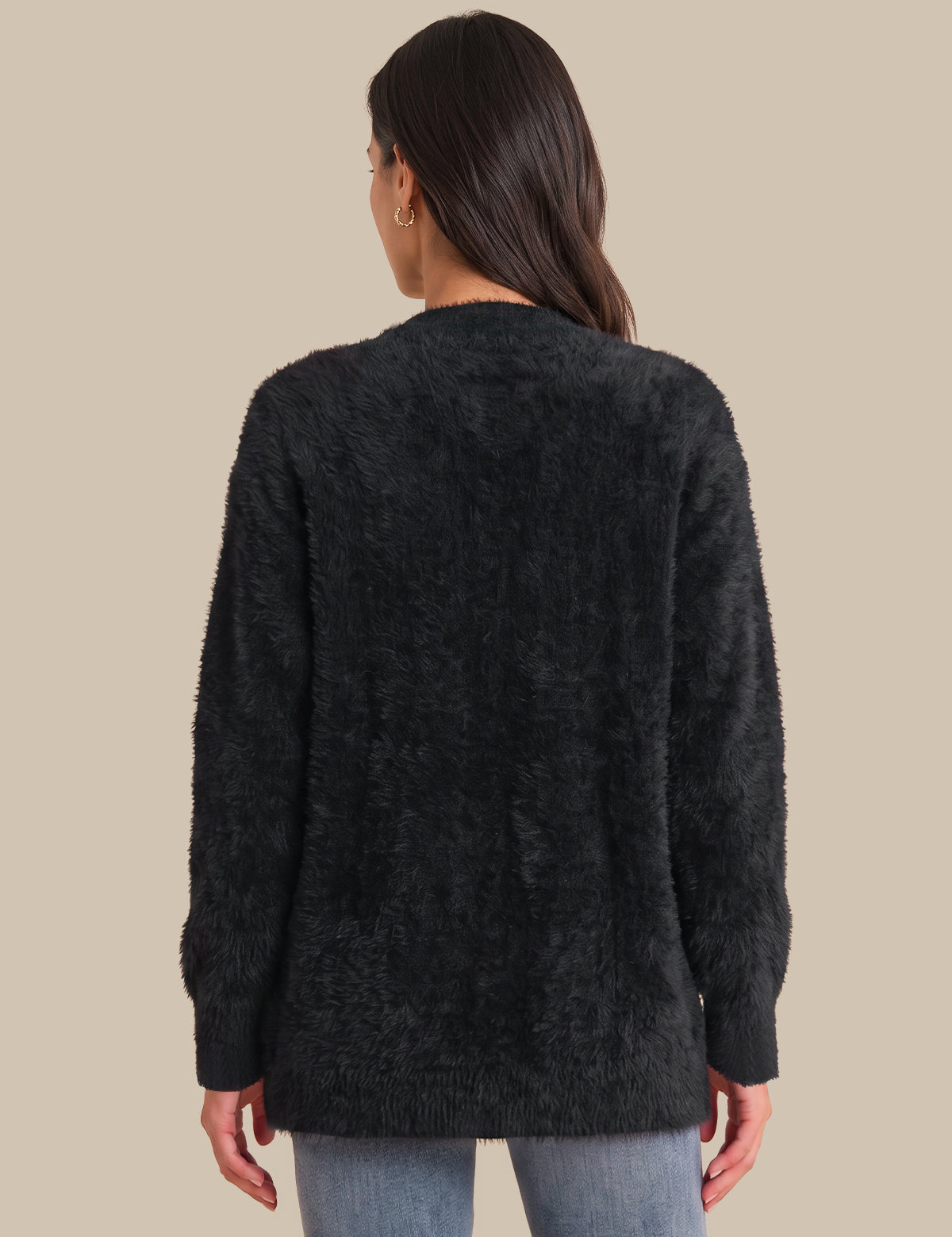 A woman viewed from the back wearing a dark charcoal fuzzy long-sleeve sweater and light blue jeans, set against a solid beige background.