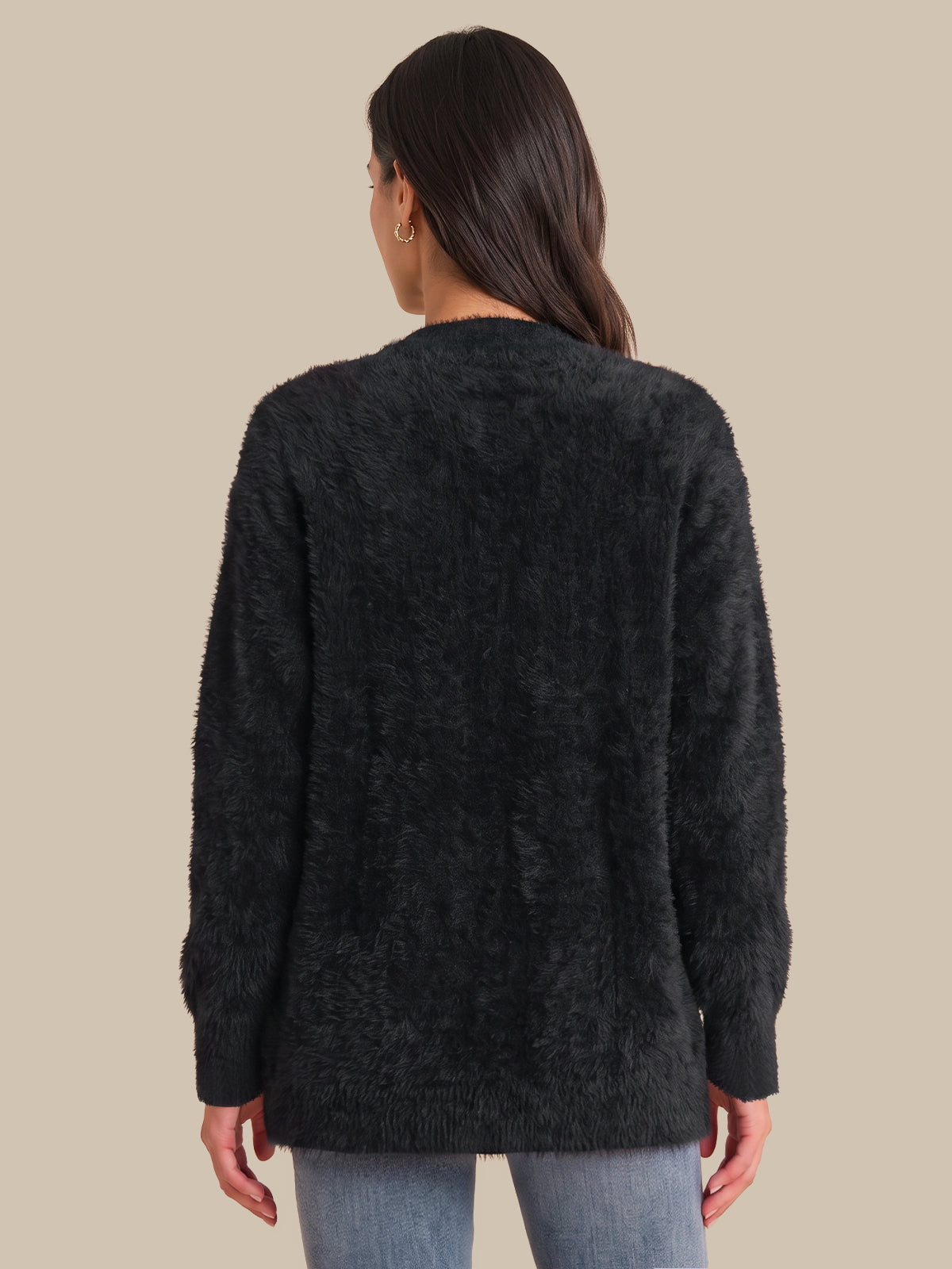 A woman viewed from the back wearing a dark charcoal fuzzy long-sleeve sweater and light blue jeans, set against a solid beige background.