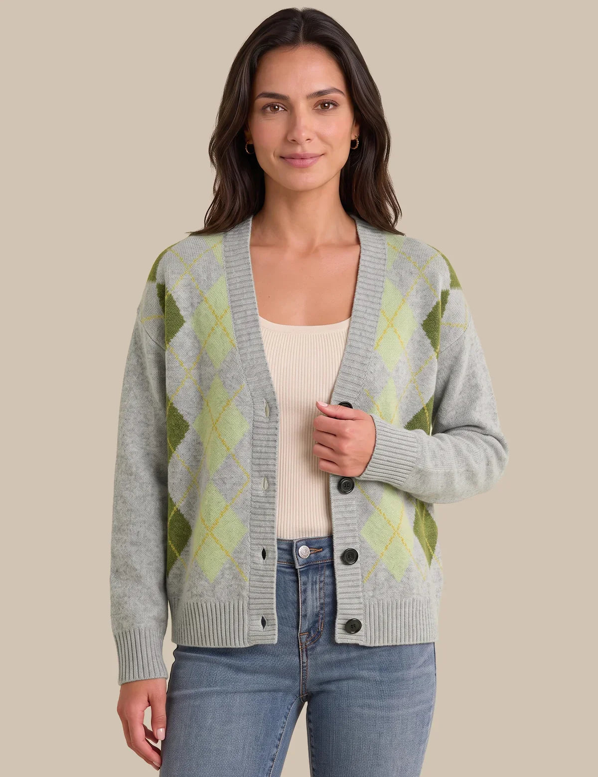 A woman modeling a light gray knit cardigan with an olive green and pale yellow argyle pattern, worn over a cream top and blue jeans against a solid beige background.