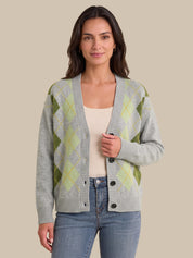 A woman modeling a light gray knit cardigan with an olive green and pale yellow argyle pattern, worn over a cream top and blue jeans against a solid beige background.