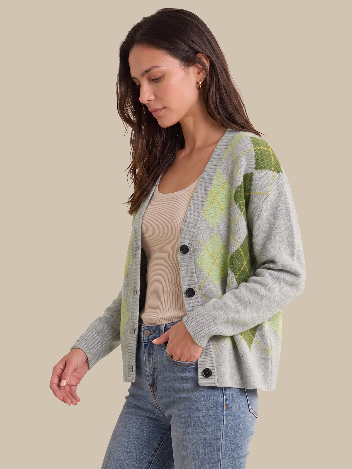 A woman wearing a light gray cardigan with green and yellow argyle patterns on the shoulders and sleeves, layered over a beige tank top and blue jeans, set against a solid beige background.