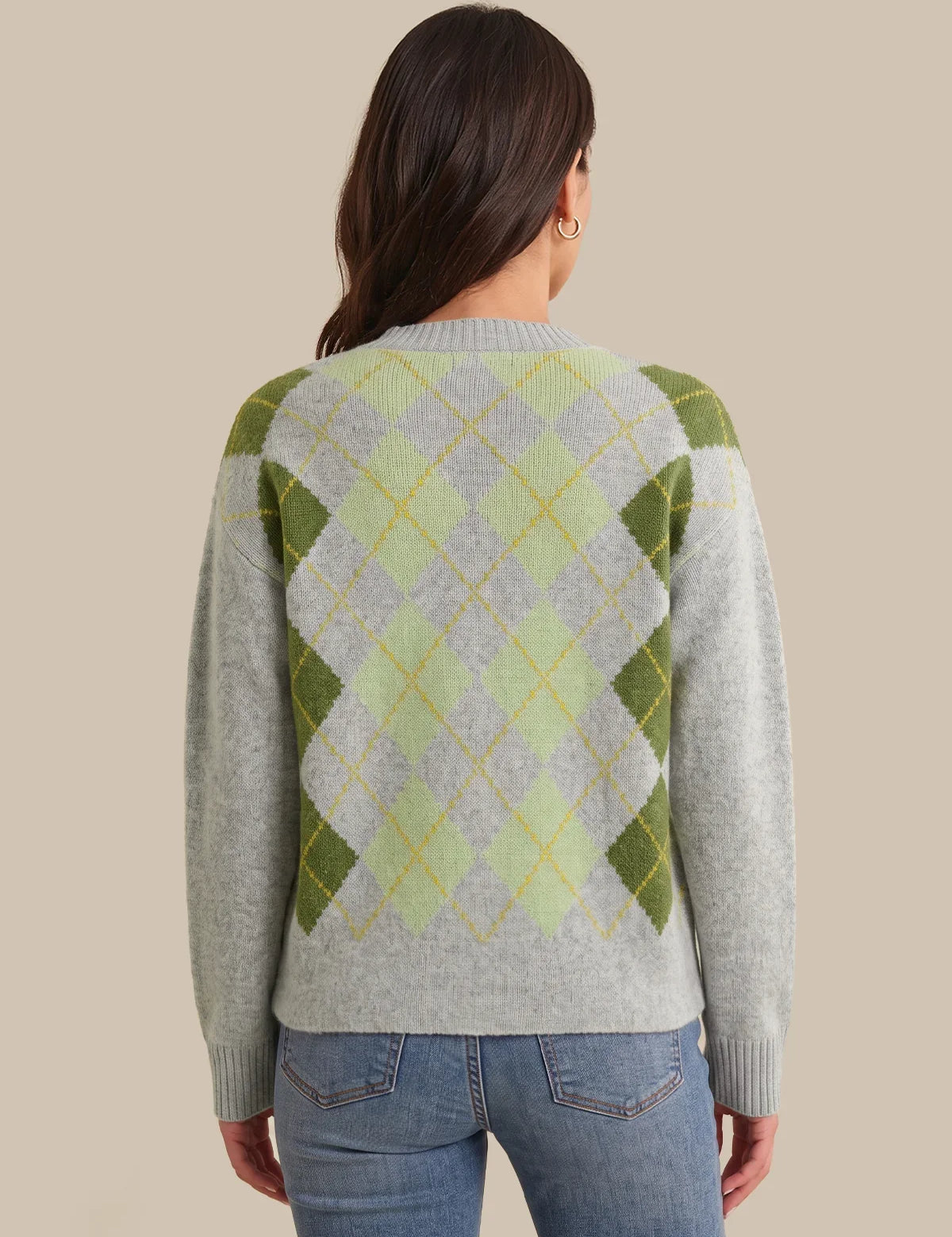 A woman viewed from the back wearing a light gray knit sweater with an argyle pattern in olive green, pale yellow, and white, paired with blue jeans against a solid beige background.