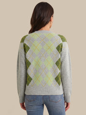 A woman viewed from the back wearing a light gray knit sweater with an argyle pattern in olive green, pale yellow, and white, paired with blue jeans against a solid beige background.