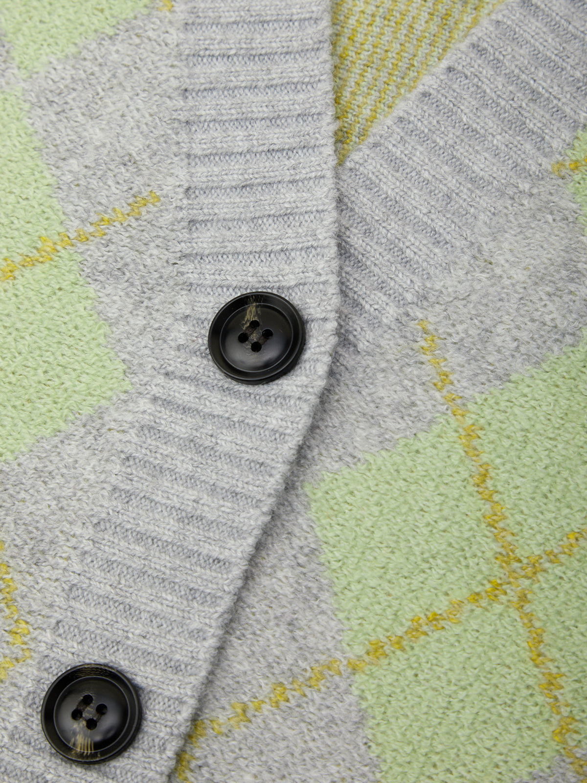 Close-up of a knit cardigan with a gray ribbed V-neck and front opening, featuring black buttons and a light green and yellow argyle pattern.