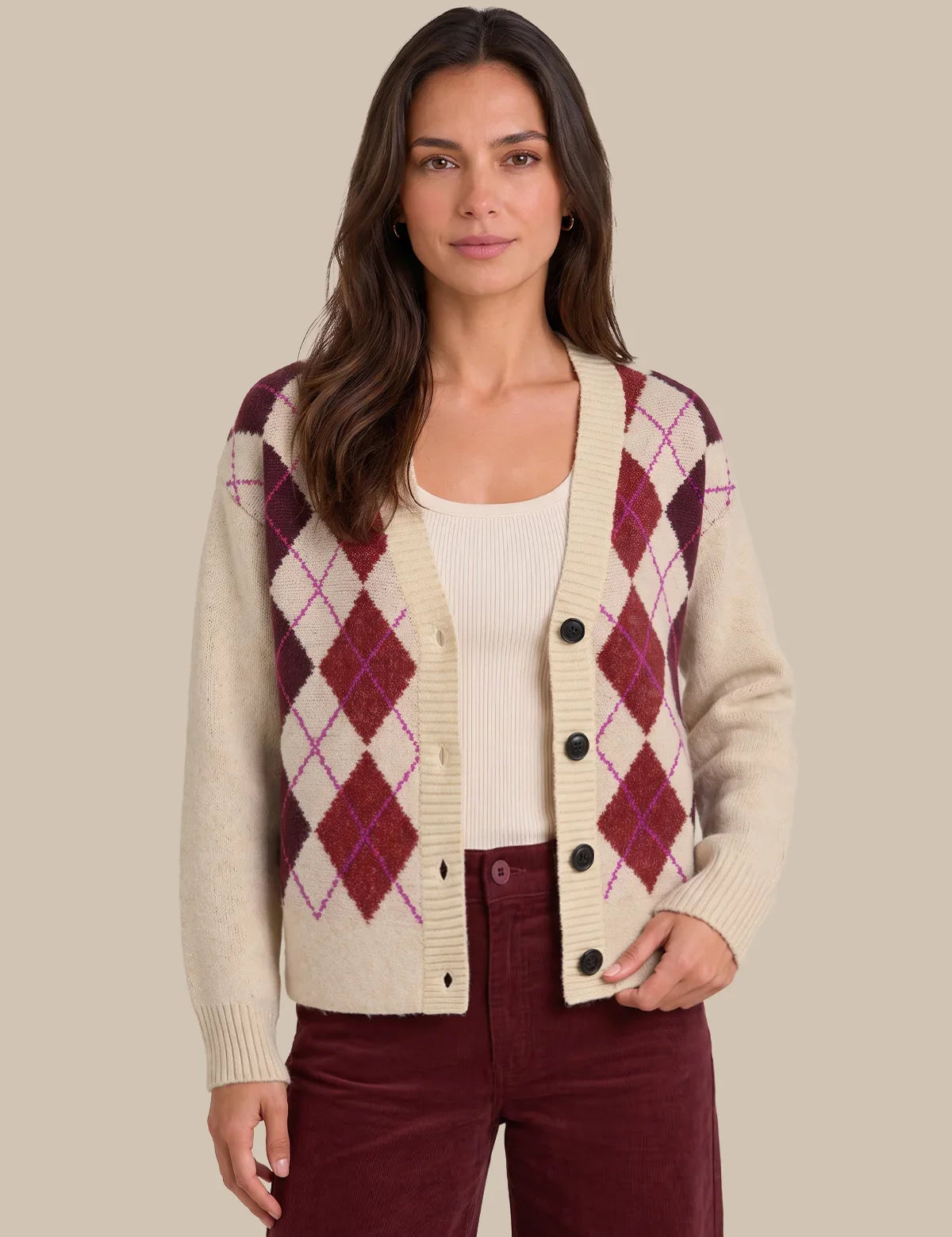 A woman modeling a cream knit cardigan with an argyle pattern in burgundy and purple, worn over a cream ribbed top and burgundy pants, against a solid beige background.
