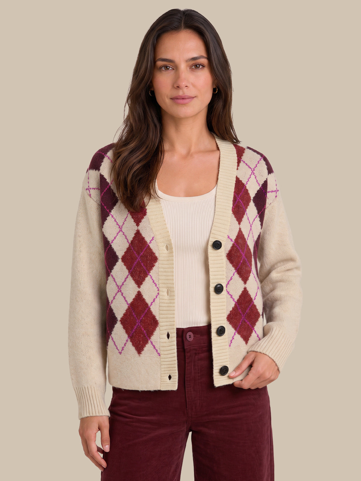 A woman modeling a cream knit cardigan with an argyle pattern in burgundy and purple, worn over a cream ribbed top and burgundy pants, against a solid beige background.