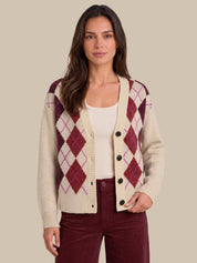 A woman modeling a cream knit cardigan with an argyle pattern in burgundy and purple, worn over a cream ribbed top and burgundy pants, against a solid beige background.