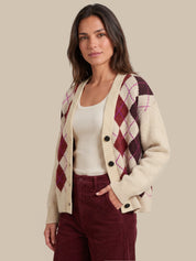 A woman modeling a cream cardigan with burgundy and magenta argyle patterns, worn over a cream tank top and burgundy pants, against a solid beige background.