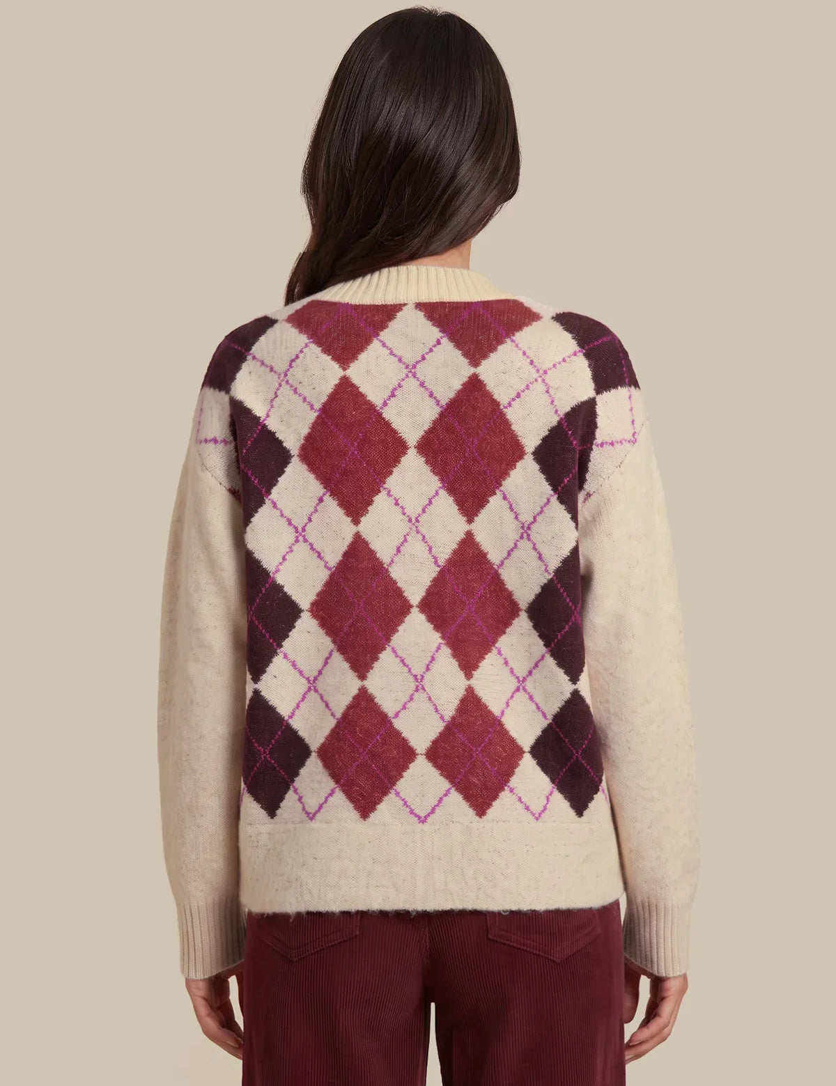 A woman shown from the back wearing a cream-colored knit sweater with a large argyle diamond pattern in burgundy, dark plum, and thin magenta lines, paired with burgundy pants, against a solid beige background.