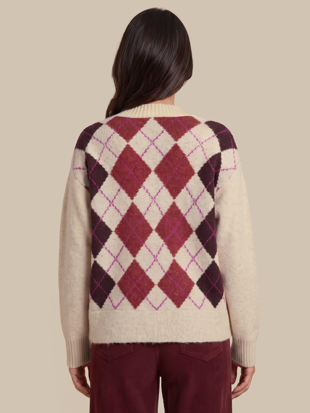 A woman shown from the back wearing a cream-colored knit sweater with a large argyle diamond pattern in burgundy, dark plum, and thin magenta lines, paired with burgundy pants, against a solid beige background.