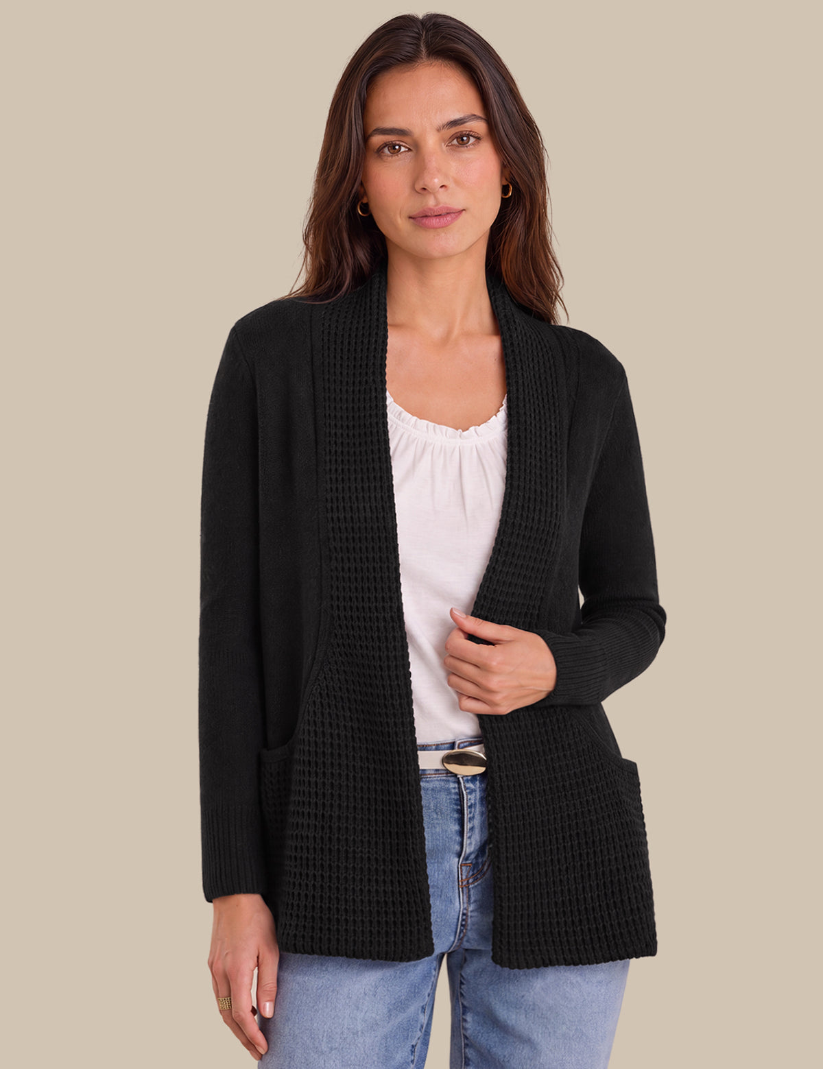 A woman modeling a black textured open-front cardigan over a white top and blue jeans, set against a solid beige background.