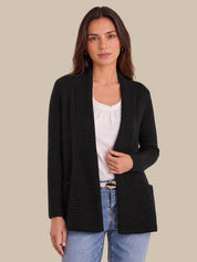 A woman modeling a black textured open-front cardigan over a white top and blue jeans, set against a solid beige background.