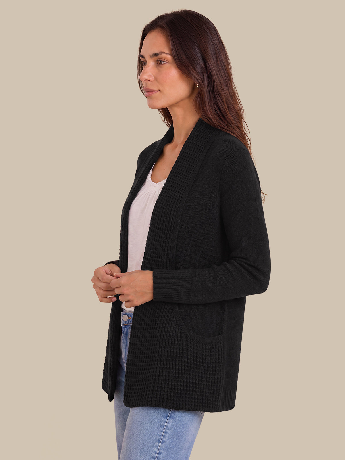 A woman wearing a black textured knit open-front cardigan over a white top and light blue jeans, shown in profile against a solid beige background.