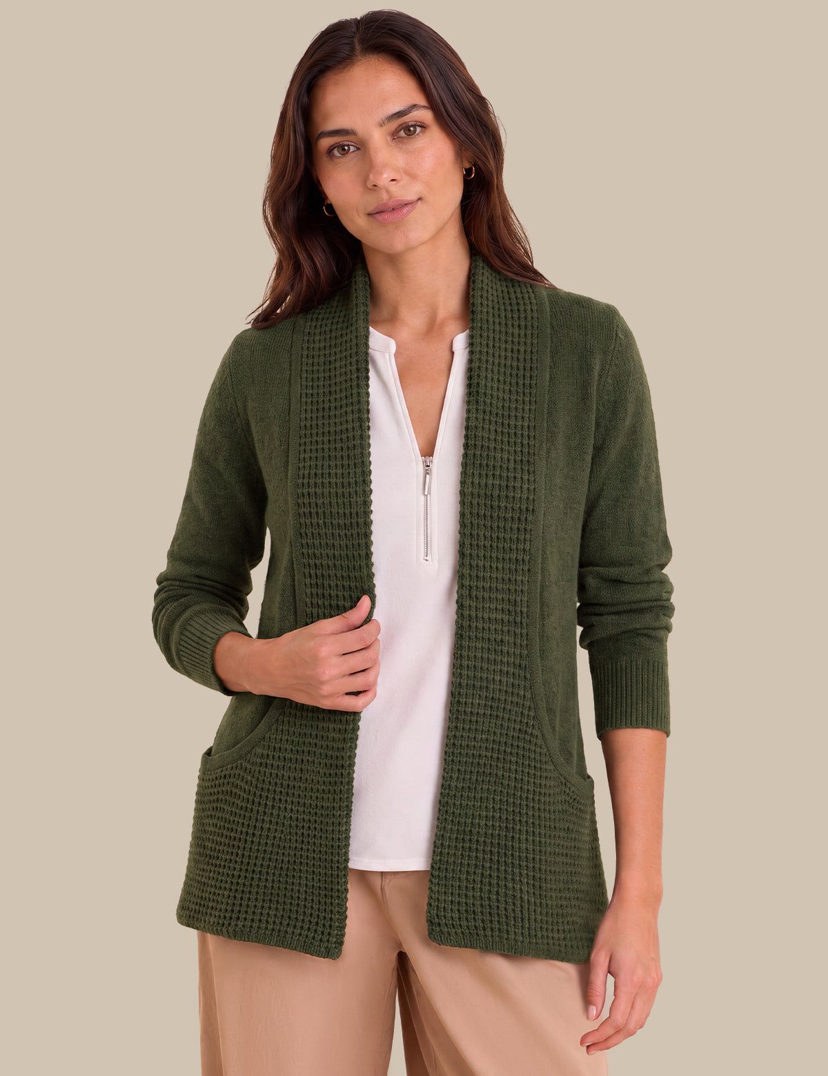 A woman wearing an olive green waffle-knit open-front cardigan over a white zip-front top and beige pants, photographed against a solid beige studio background.