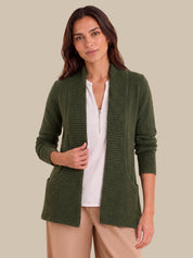 A woman wearing an olive green waffle-knit open-front cardigan over a white zip-front top and beige pants, photographed against a solid beige studio background.