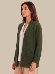 A woman wearing an olive green open-front knit cardigan with textured vertical panels and side pockets, layered over a white top and beige pants, against a solid beige background.