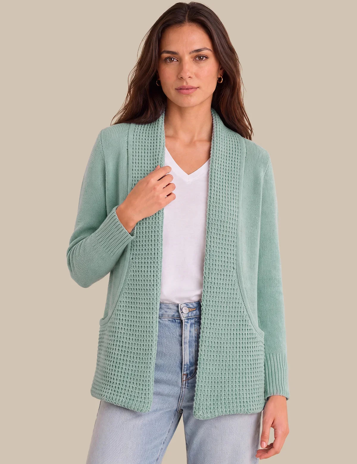 A woman modeling a sage green open-front waffle-knit cardigan over a white V-neck top and light-wash blue jeans, set against a solid beige background.