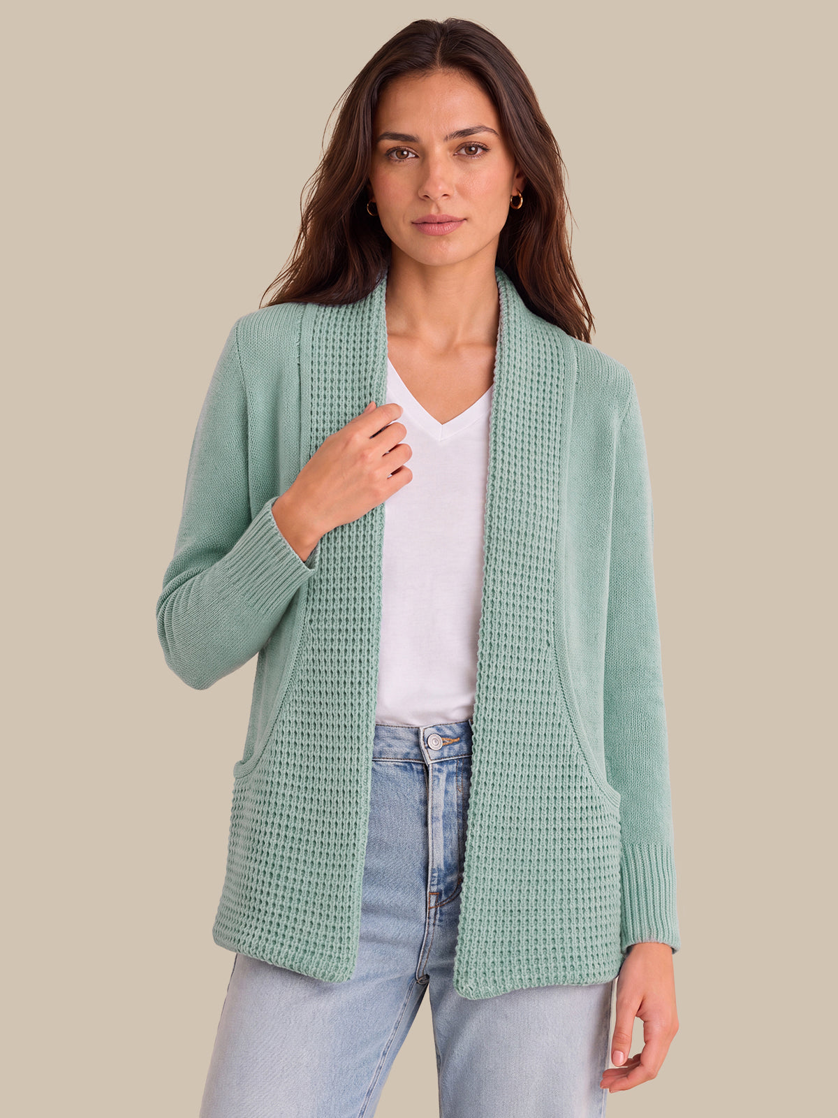 A woman modeling a sage green open-front waffle-knit cardigan over a white V-neck top and light-wash blue jeans, set against a solid beige background.