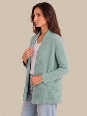 A woman wearing a sage green open-front knit cardigan with a waffle texture over a white top and light blue jeans, set against a solid beige background.