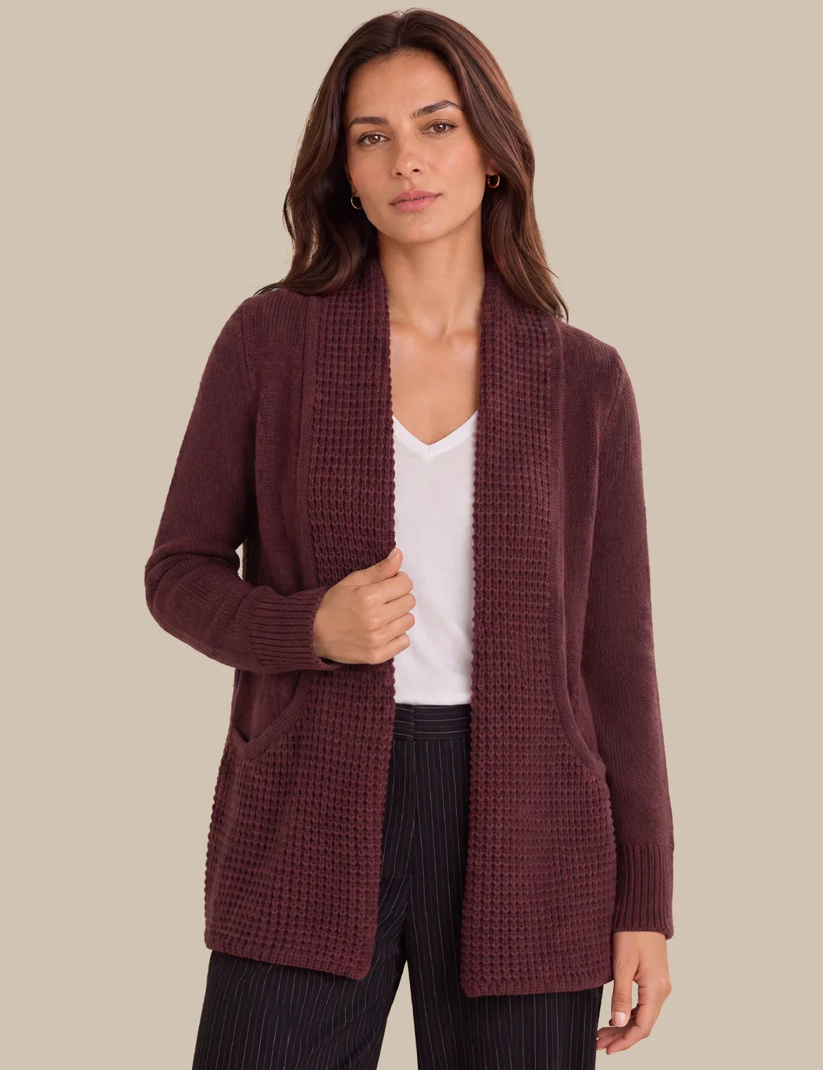 A woman modeling a textured, open-front cardigan in a deep burgundy color, worn over a white top and dark pinstriped trousers, set against a solid beige background.