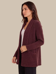 A woman wearing a textured, open-front cardigan in a deep burgundy or plum color, layered over a white top and dark pinstriped pants, set against a solid beige background.