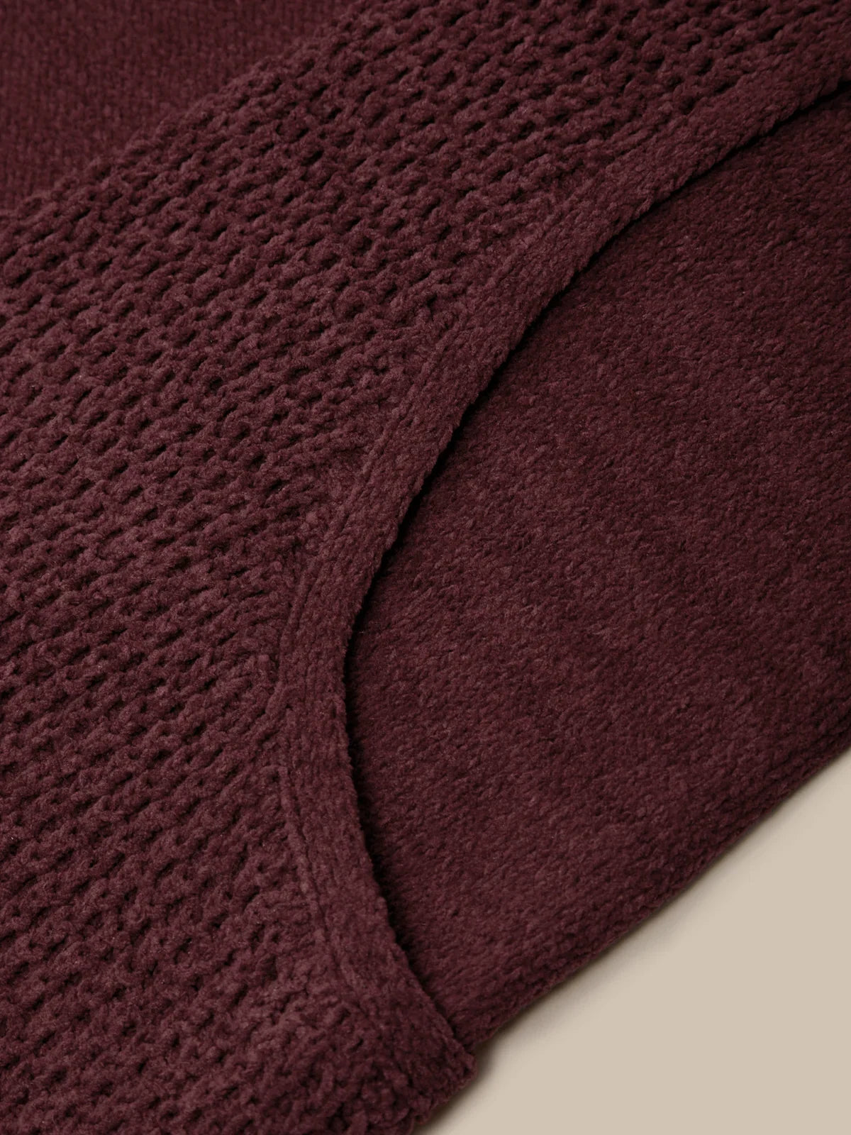 Close-up of a textured knit garment in a deep burgundy or plum color, showing the waffle-knit pattern and curved neckline against a light beige background.