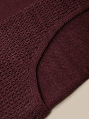 Close-up of a textured knit garment in a deep burgundy or plum color, showing the waffle-knit pattern and curved neckline against a light beige background.