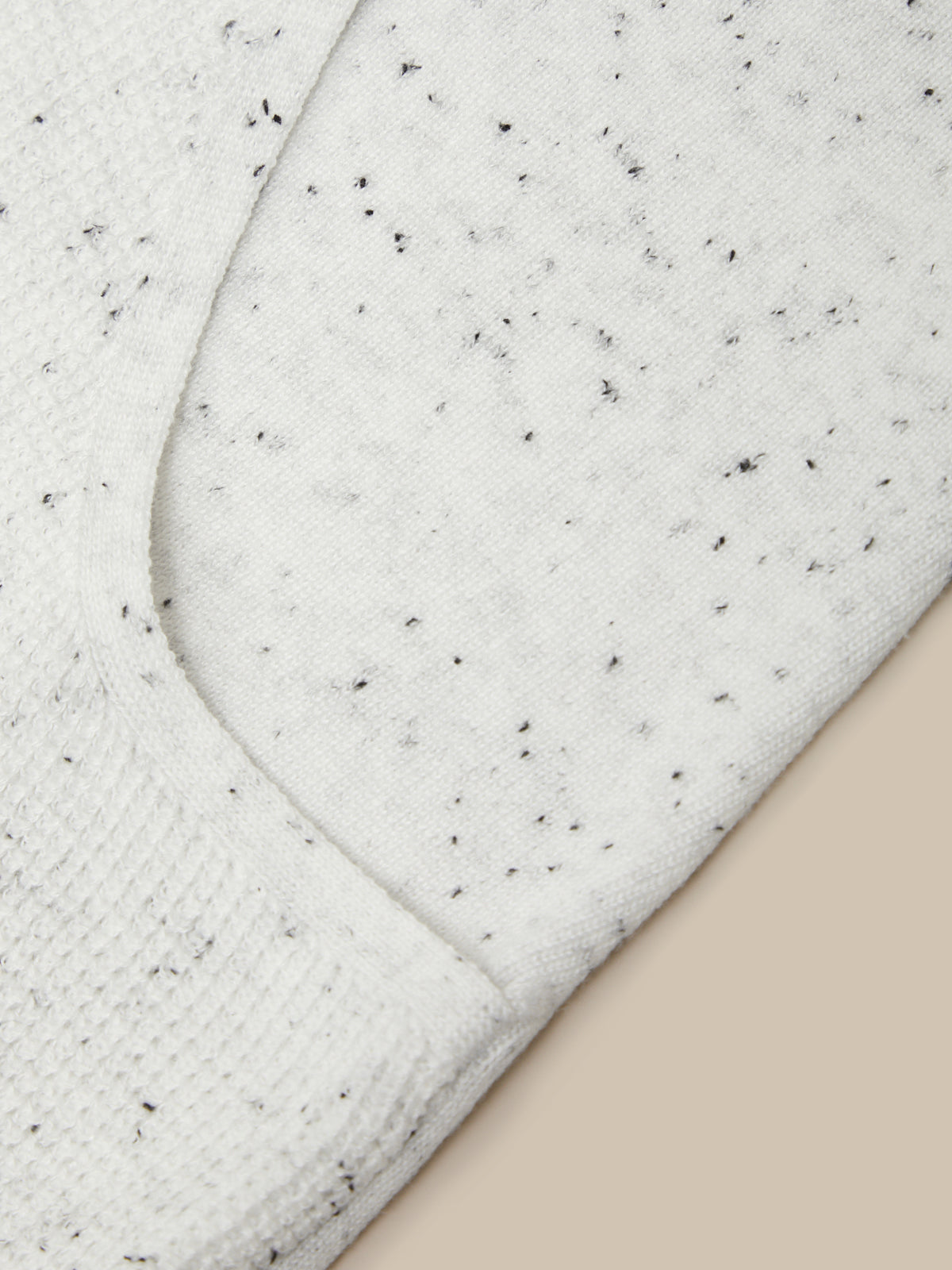 Close-up of a white or off-white knit fabric with small black speckles, showing a curved seam and ribbed texture against a light beige background.