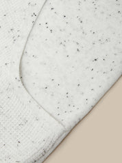 Close-up of a white or off-white knit fabric with small black speckles, showing a curved seam and ribbed texture against a light beige background.