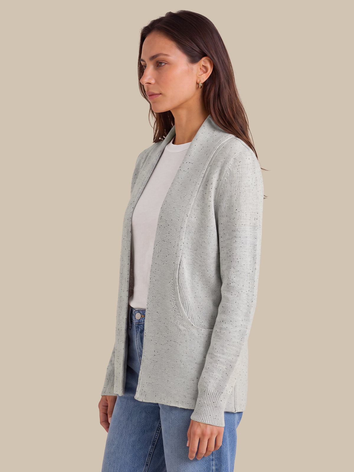 A woman wearing a light gray open-front cardigan over a white top and blue jeans, shown in profile against a solid beige background.