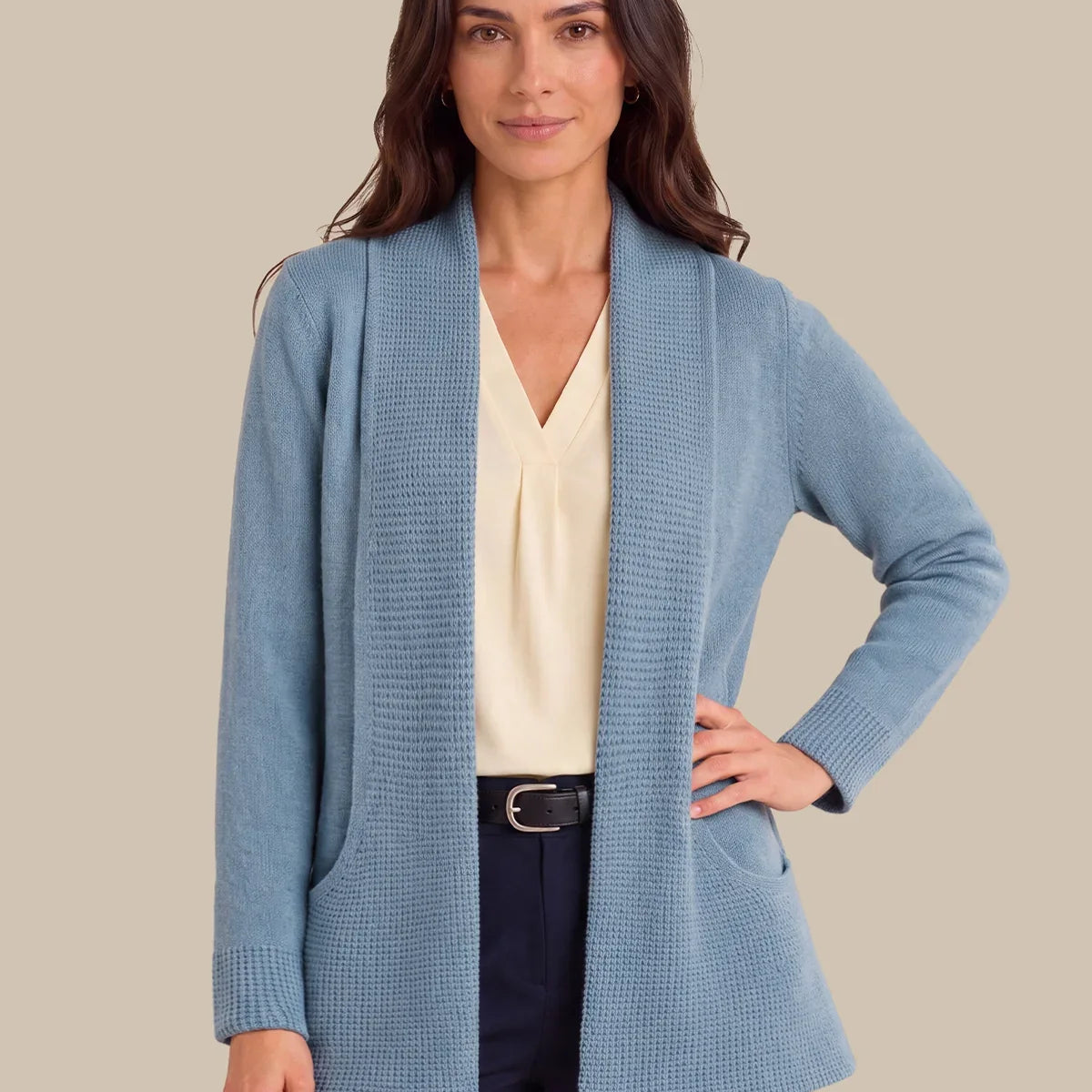 A woman modeling a light blue textured knit shawl-collar cardigan over a cream blouse and navy trousers, against a solid beige background.
