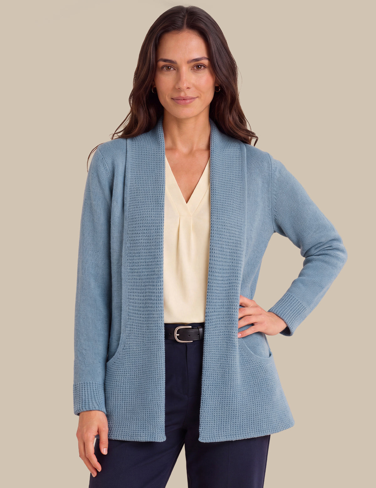 A woman modeling a light blue textured knit shawl-collar cardigan over a cream blouse and navy trousers, against a solid beige background.