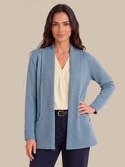 A woman modeling a light blue textured knit shawl-collar cardigan over a cream blouse and navy trousers, against a solid beige background.