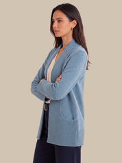 A woman wearing a light blue ribbed knit cardigan with front patch pockets, layered over a cream top and dark trousers, standing against a solid beige background.