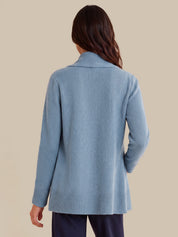 A woman viewed from the back wearing a light blue knit cardigan with a ribbed collar, cuffs, and hem, paired with dark navy pants, against a solid beige background.