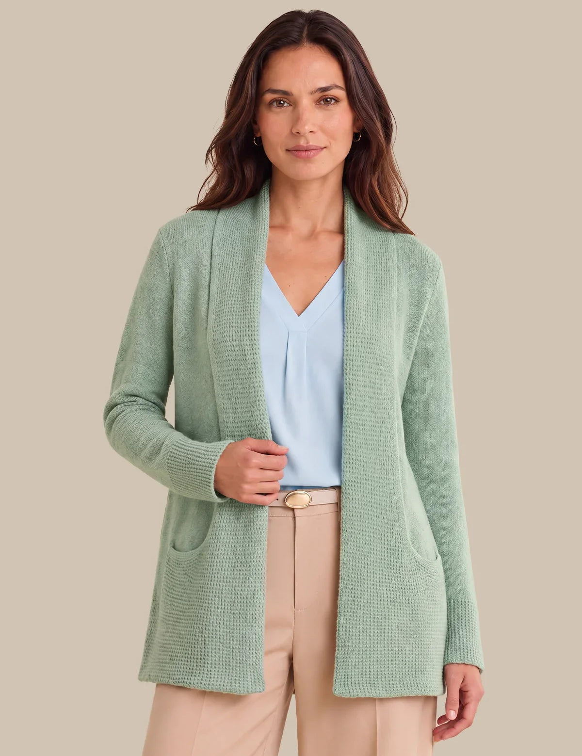 A woman modeling a light sage green open-front knit cardigan over a light blue blouse and beige trousers, set against a solid beige background.