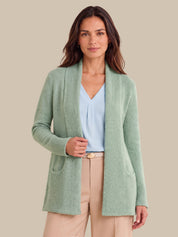 A woman modeling a light sage green open-front knit cardigan over a light blue blouse and beige trousers, set against a solid beige background.