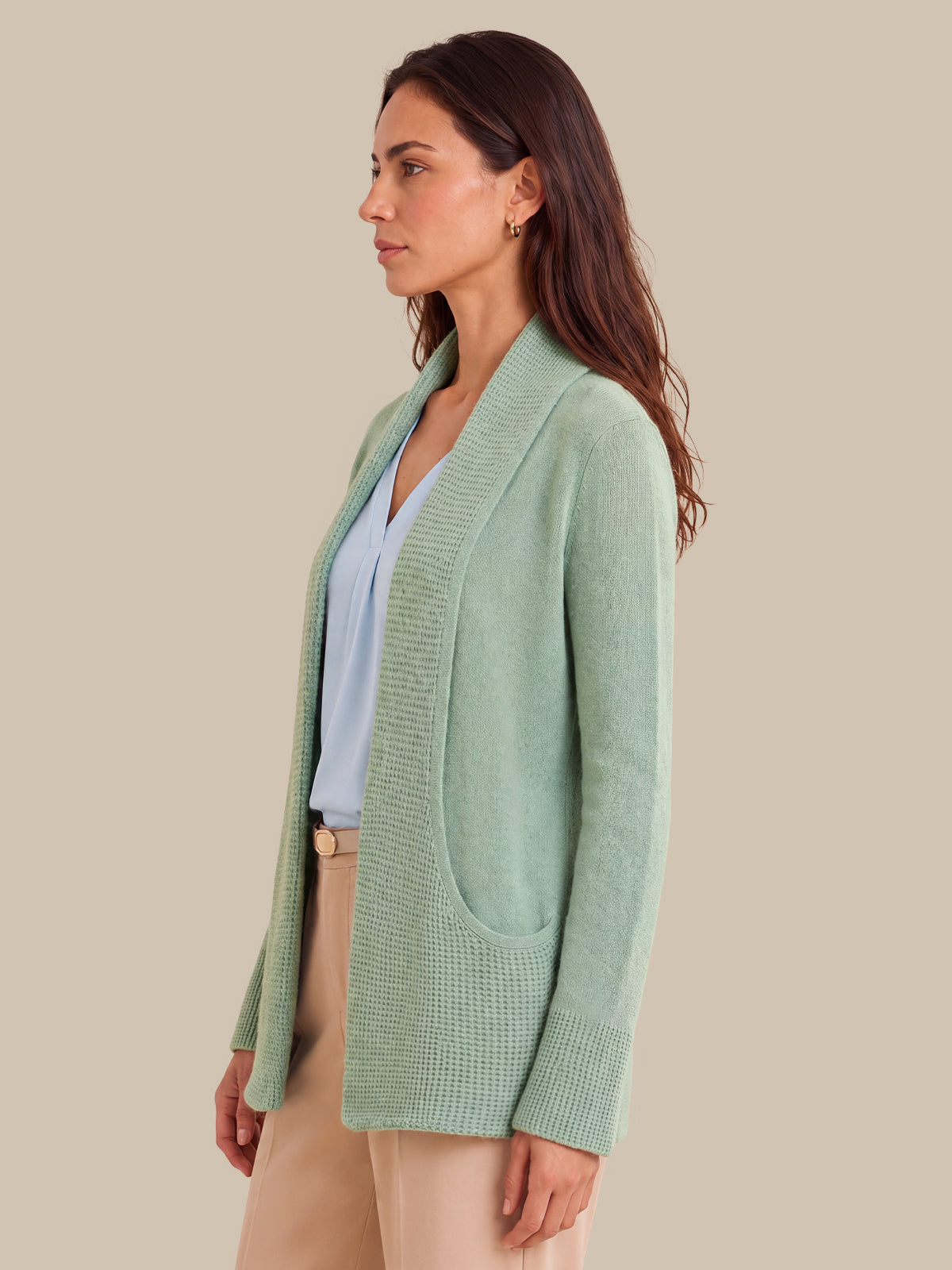 A woman shown in side profile wearing a light sage green open-front knit cardigan over a pale blue top and beige pants, against a plain warm beige studio background.
