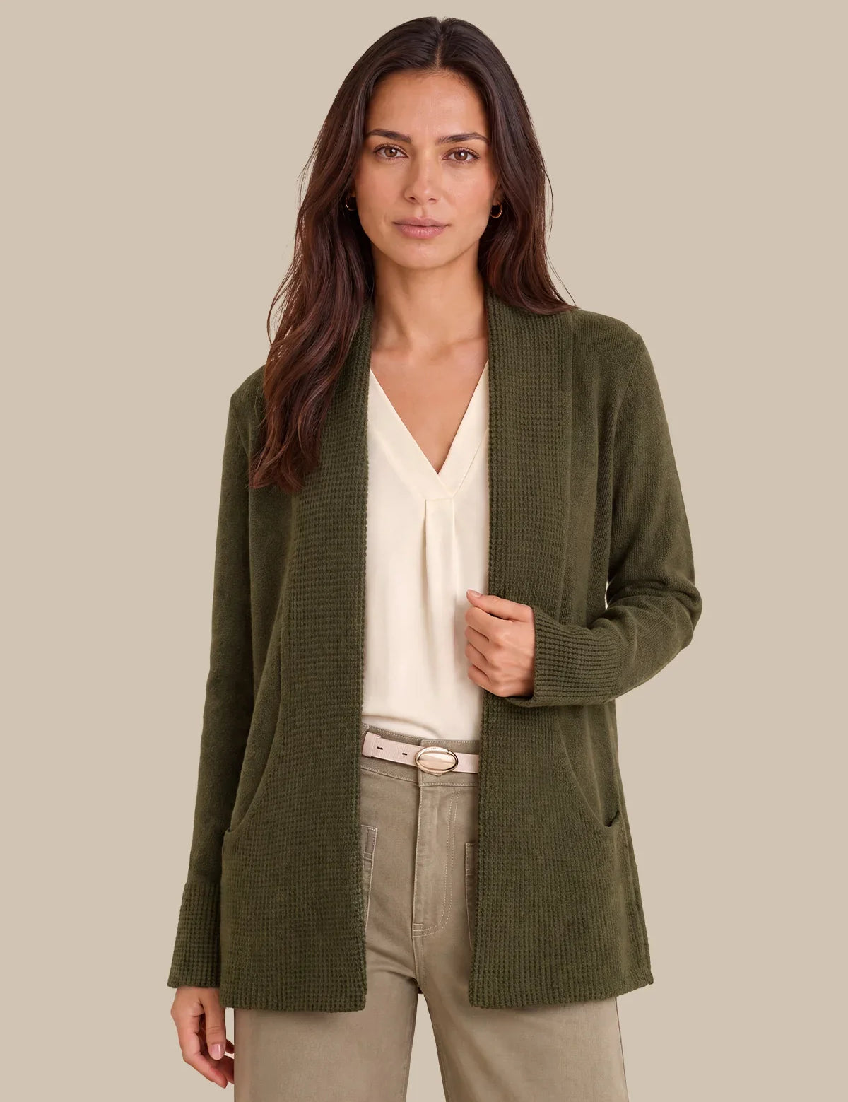 A woman modeling an olive green ribbed knit cardigan over a cream blouse and beige trousers, set against a solid beige background.