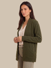 A woman wearing an olive green open-front knit cardigan over a cream blouse and beige trousers, photographed against a plain warm beige studio background.