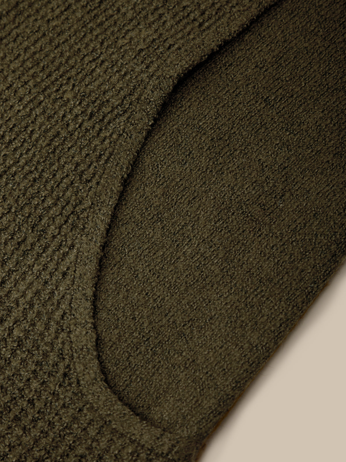 Close-up of a dark olive green knit fabric with a ribbed texture and a curved seam detail, set against a light beige background.