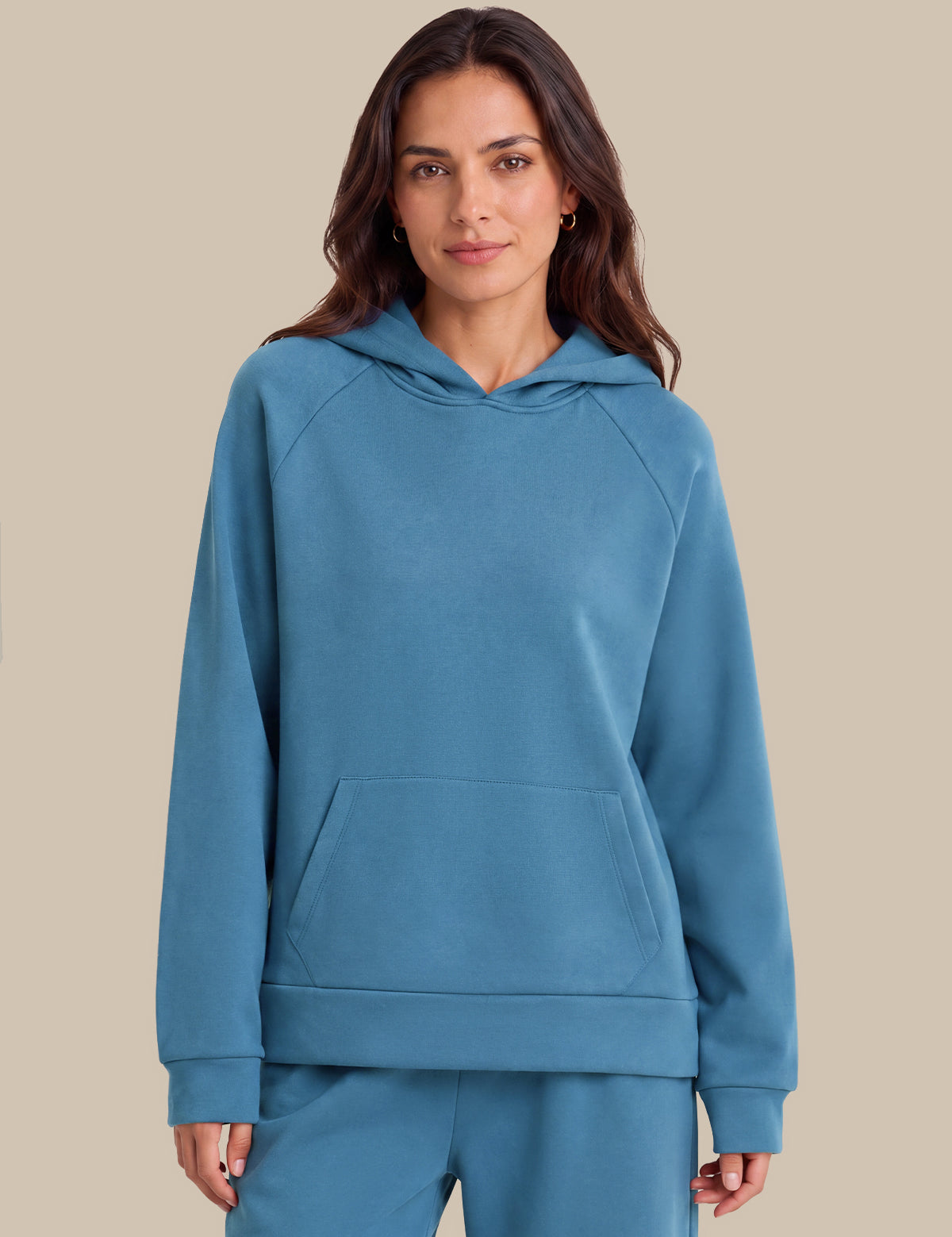 A woman modeling a matching slate blue loungewear set, featuring a pullover hoodie with a kangaroo pocket and matching sweatpants, set against a solid beige background.