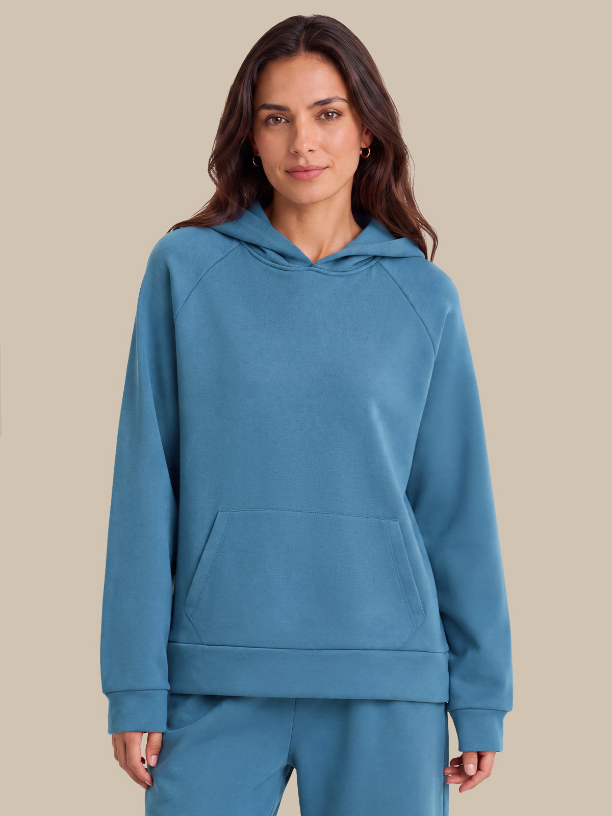 A woman modeling a matching slate blue loungewear set, featuring a pullover hoodie with a kangaroo pocket and matching sweatpants, set against a solid beige background.